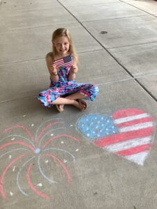 A little girl holds an American flag above a chalk drawing of a firework and a heart